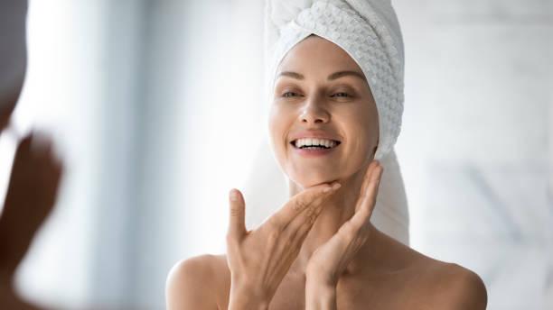 Happy lady look in bathroom mirror touching healthy face skin Happy confident young lady with towel on head look in bathroom mirror touching moisturized soft healthy sensitive skin doing morning hydration spa beauty routine, facial skincare treatment concept hydrated skin stock pictures, royalty-free photos & images