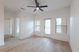 Bright, empty apartment interior with hardwood floors, ceiling fan, and doors leading to a balcony.