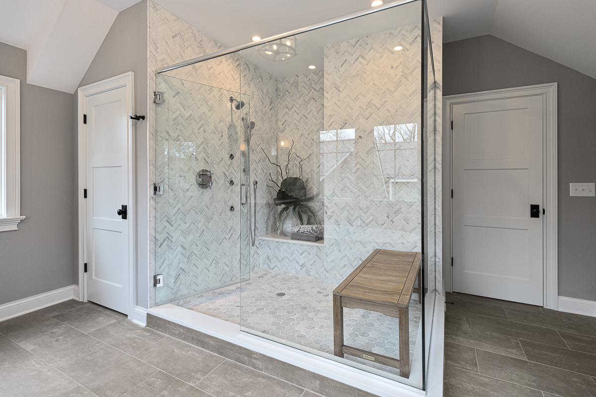 A large walk in shower with multiple shower heads, full glass surround, honeycomb and chevron patterned tiles. Featuring a large recessed nook and teak shower seat.