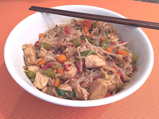 Chicken pieces with bean sprouts and vegetables in a bowl with chop sticks on an orange mat.