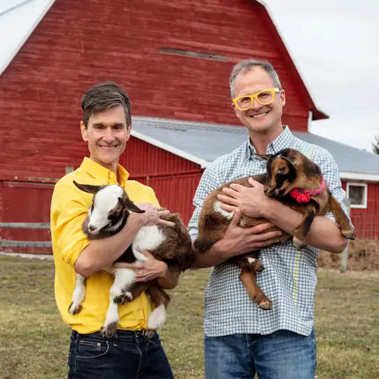 Los fundadores de Beekman 1802, Josh Kilmer-Purcell y Brent Ridge, sonríen a la cámara mientras posan con cabras en un establo.