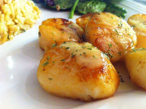 A few sautéed scallops sitting on a white plate topped with sauce with rice and green beans in the background.