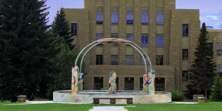 Artists redition of a piece of art with 4 pillars with rainbow colors that meet in an arch over a central pillar. The art is showcased in front of the University of Wyoming College of Arts & Sciences.