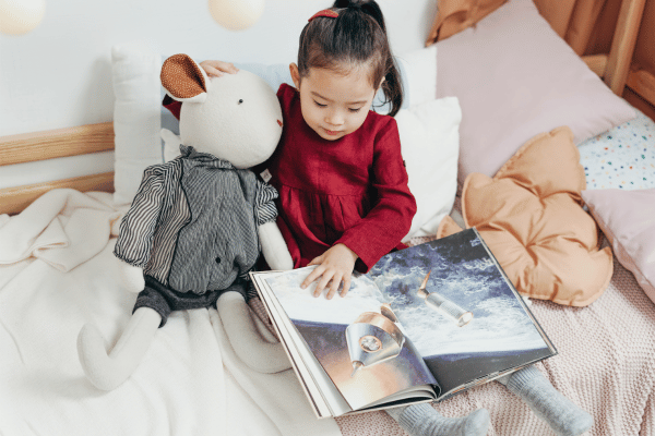 Young girl sitting on a soft cushion reading a picture book and holding a stuffed animal.