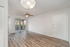 Apartment entry door open to a living area with wood plank flooring, tan walls, white trim, a ceiling fan, and patio doors with vertical blinds 