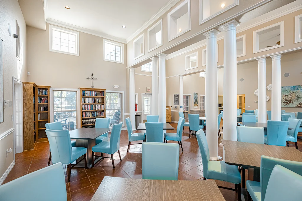 Clubhouse seating area with tables and blue chairs, bookshelves, tall pillars, light walls, and square tile flooring