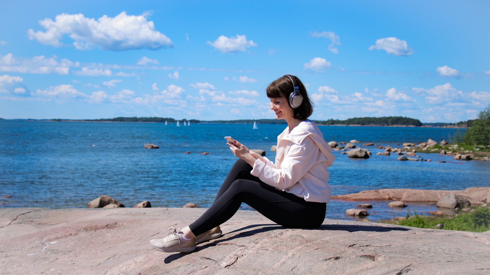 Nainen istuu meren rannalla kalliolla, kuulokkeet korvilla, katsoo puhelinta ja hymyilee.