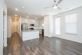 Open concept living room with wood-inspired flooring, white walls, a large window, ceiling fan, and access to the kitchen