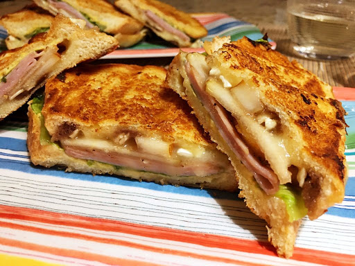 A sandwich on a multi-color plate with a glass of white wine in the background.