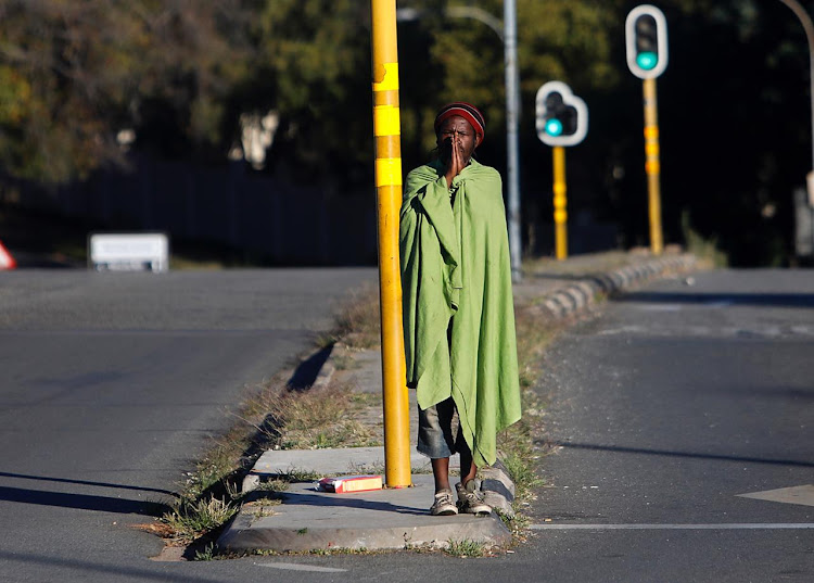 A man clad in a blanket stands on an extremely quiet street corner, in Jozi.