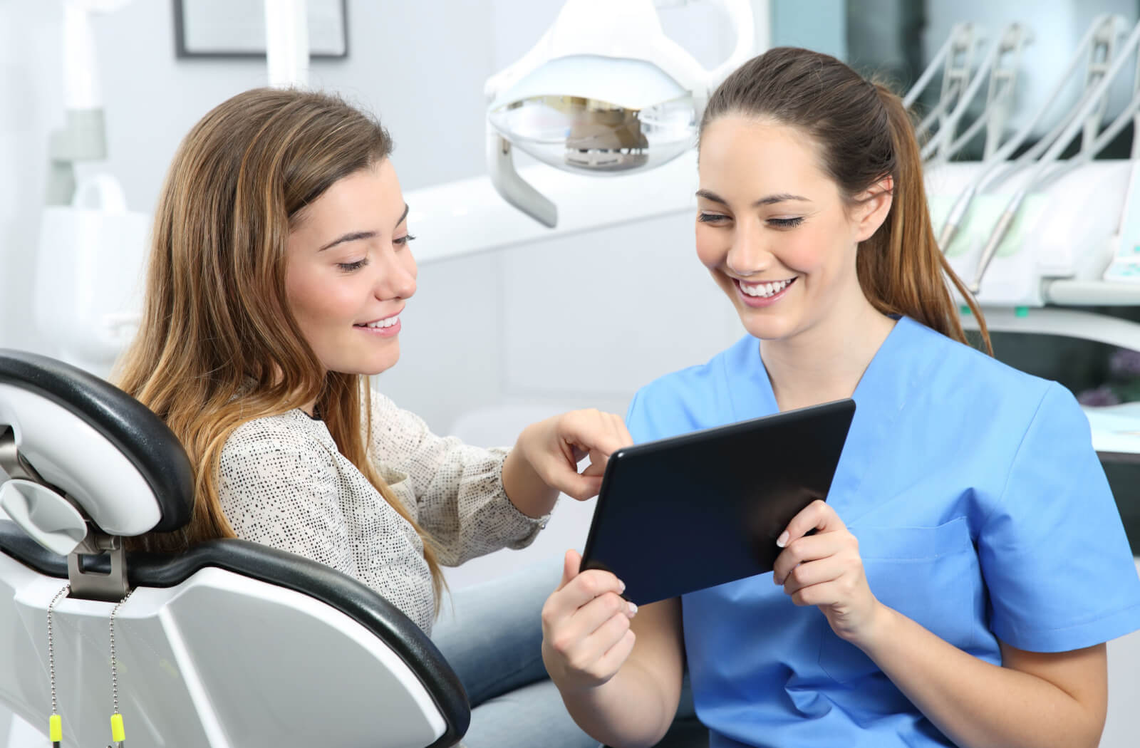 A female dentist in blue scrubs and a woman sitting in a dentist's chair looking at a tablet and smiling.