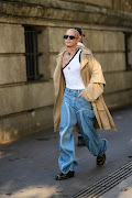 A guest wears a white with black borders Bon Bom asymmetric tank-top, a beige oversized coat, blue faded denim large ruffled pants and black shiny leather shoes outside Rick Owens, during Paris Fashion Week - Womenswear Spring/Summer 2023. 
