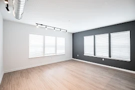 Living room with six windows with blinds, dark grey accent wall, track lighting, exposed ductwork, and light plank flooring