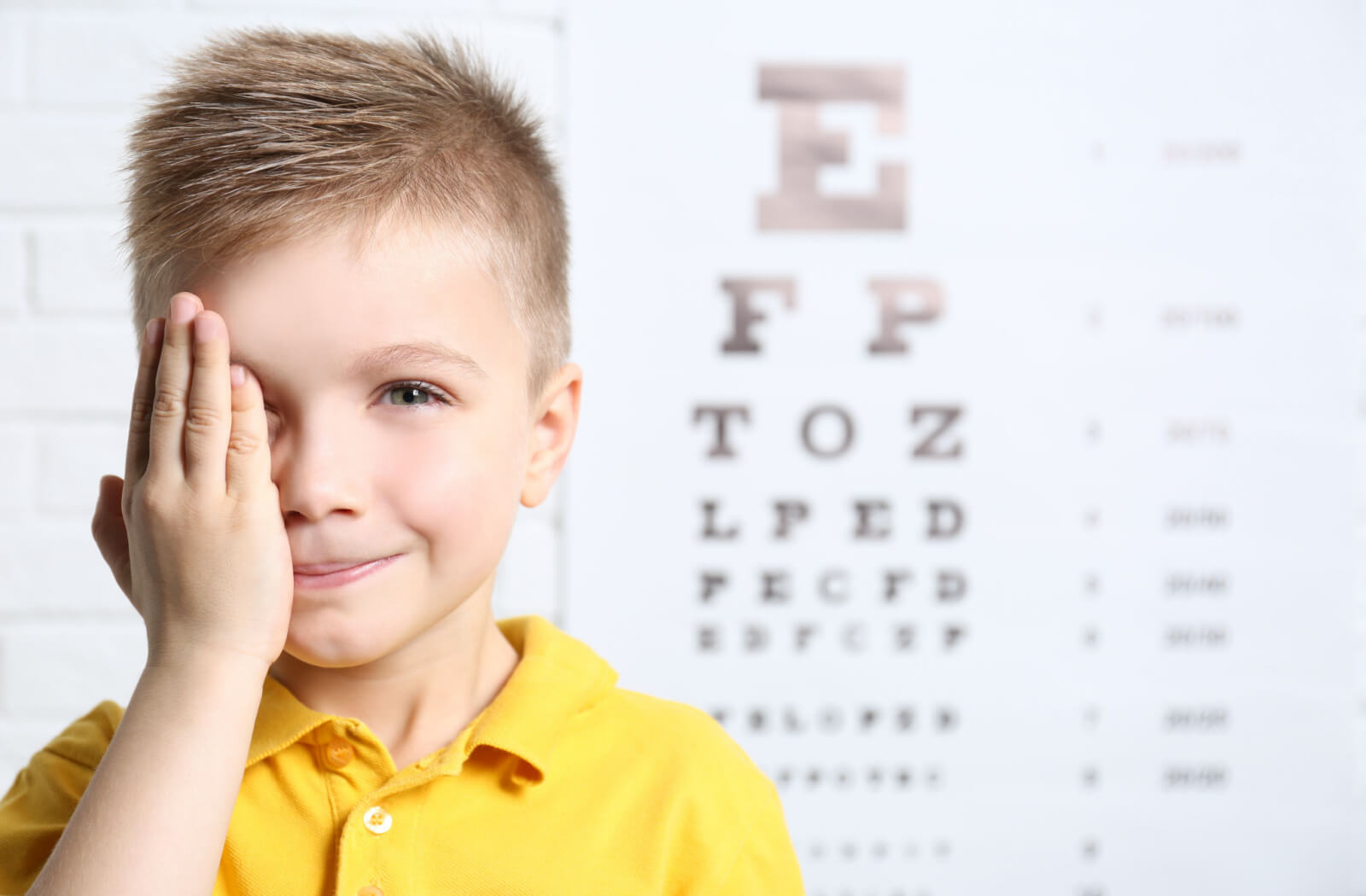 A boy in a yellow collared shirt is covering his right eye with his right hand while he is undergoing a Snellen chart exam.