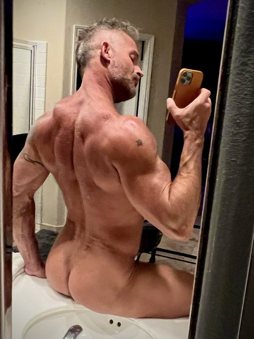 Greg Dixxon sitting on the bathroom sink naked showing off his muscled back and ass while taking an iphone mirror selfie