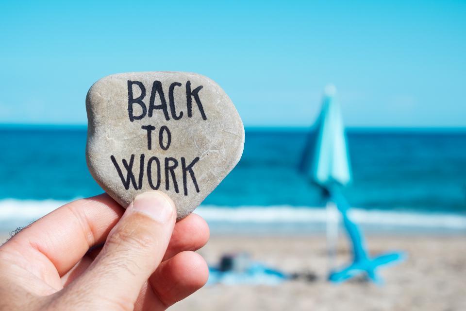 A hand holds a smooth shell that says Back to Work on it. In the background is the Ocean. 