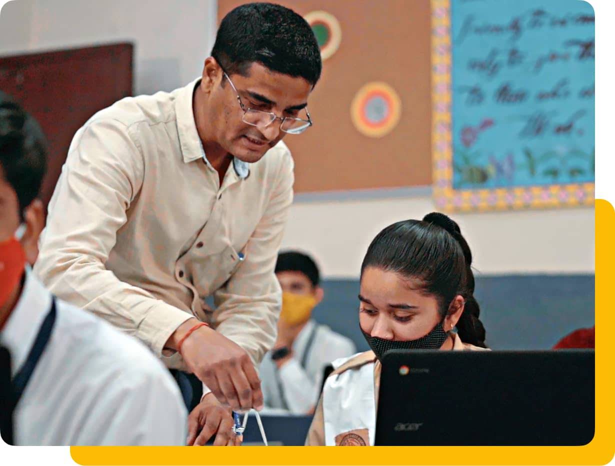 In a classroom, a female student works on her Chromebook at her desk. A male teacher leans over to help.