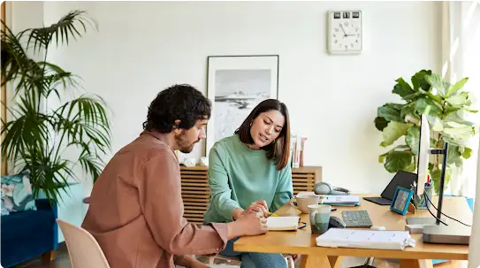 Um homem e uma mulher sentados à mesa no escritório de casa, lendo anotações juntos. A mulher aponta para uma página e o homem observa.