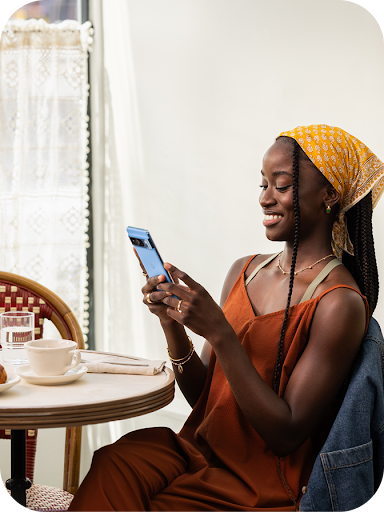 En person med långa flätor som har en gul huvudduk och en rostfärgad klänning sitter vid ett bord och ler medan hen använder en smartphone. Platsen verkar vara ett ljust kafé eller en restaurang.