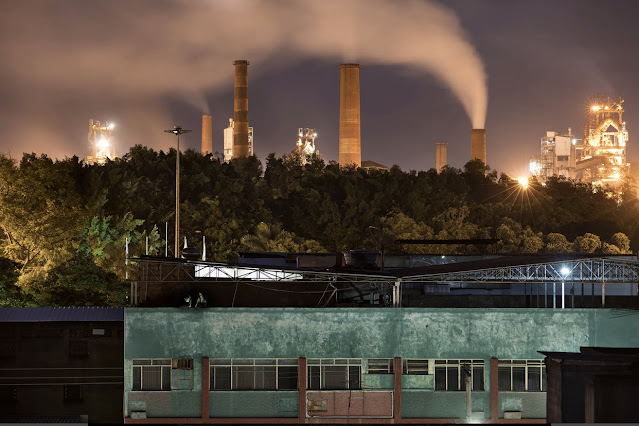 A imagem mostra um complexo residencial cinza escuro. Ao fundo, é possível ver o céu roxo com grandes nuvens de fumaça proveniente da usina da cidade e grandes chaminés.