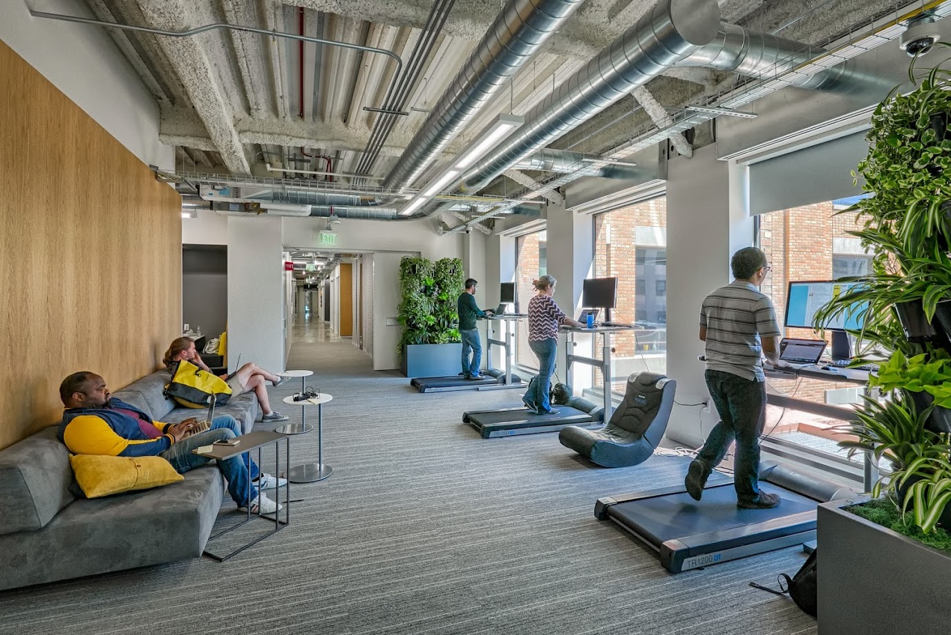 An office with floor to ceiling windows. Treadmills face the window. A few people are walking on the treadmills.