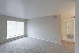 Living Room with neutral walls and carpet with light trim & a large window with blinds on the left & a hallway on the right.