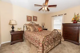 Cozy bedroom with queen bed, patterned bedding, dresser, side table, lamp, and ceiling fan in an apartment setting.