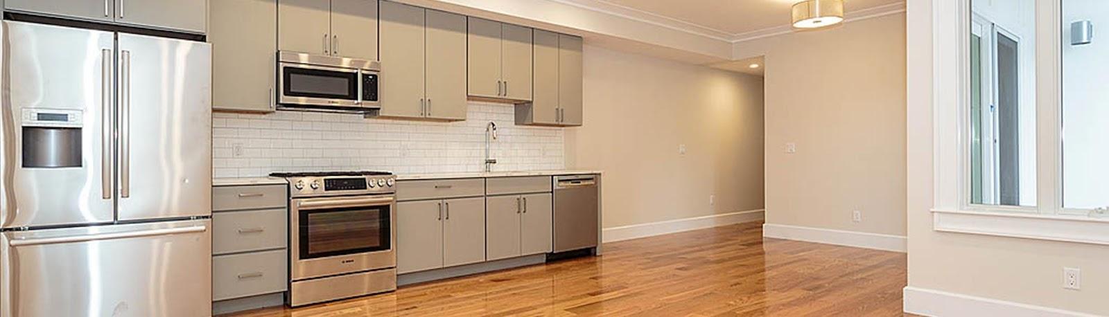 A white, unfurnished kitchen with gray cabinets, silver appliances, and hardwood floors.