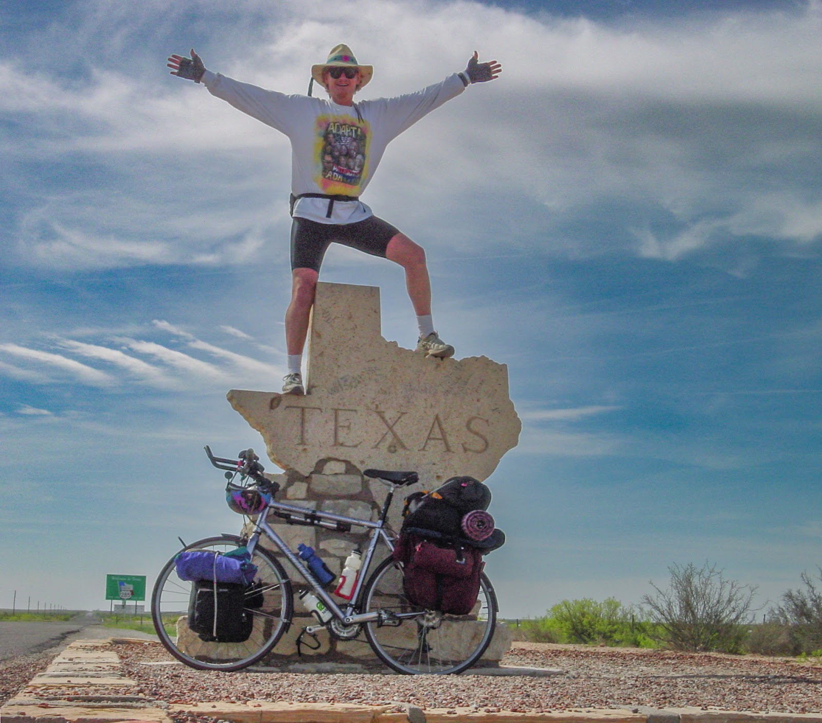 Man stands atop the Texas state line marker in the shape of the state, bicycle leans on the sign below. Blue sky with clouds in open country. 