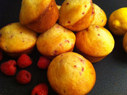 Seven to eight muffins with raspberries on a black table mat with a lemon in the background.