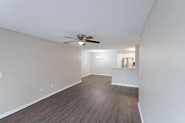 Livingroom with grey wood flooring & grey walls. Dark colored ceiling fan overhead with a dining area and kitchen in the back