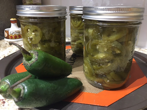 Three jars of pickled jalapenos with three jalapeno peppers on a baking sheet lined with a silicone mat.