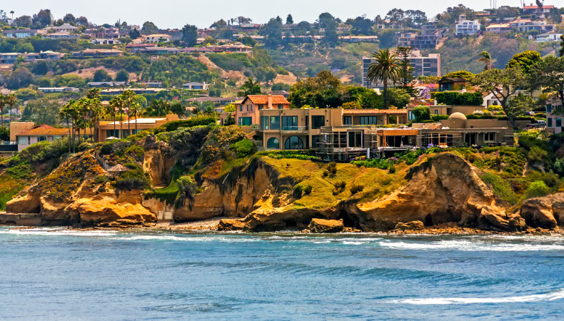 A beautiful view of the La Jolla neighborhood in San Diego. Green rocky cliffs and sandy beaches line the coast with large beautiful homes rising up behind them.