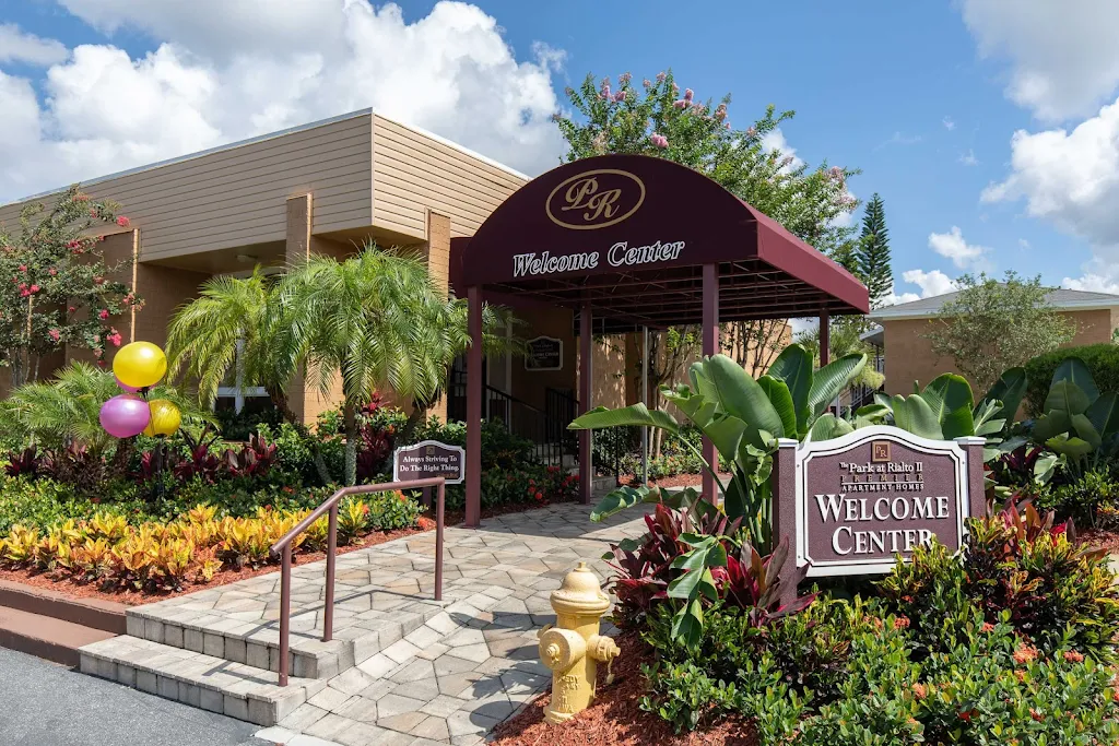 Front entrance of an apartment complex welcome center with a maroon awning, colorful balloons, and landscaped garden.