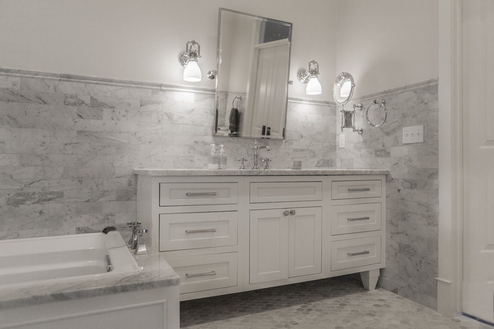 White and gray guest bathroom featuring a narrow wall mounted single sink vanity with drawers flanking either side of the under sink cabinet.  An adjustable large rectangular mirror hangs above the  sink with upside down tulip shaped sconces on either side.