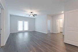 Empty apartment interior with gray walls, wood floor, ceiling fan, windows, and doors to other rooms.