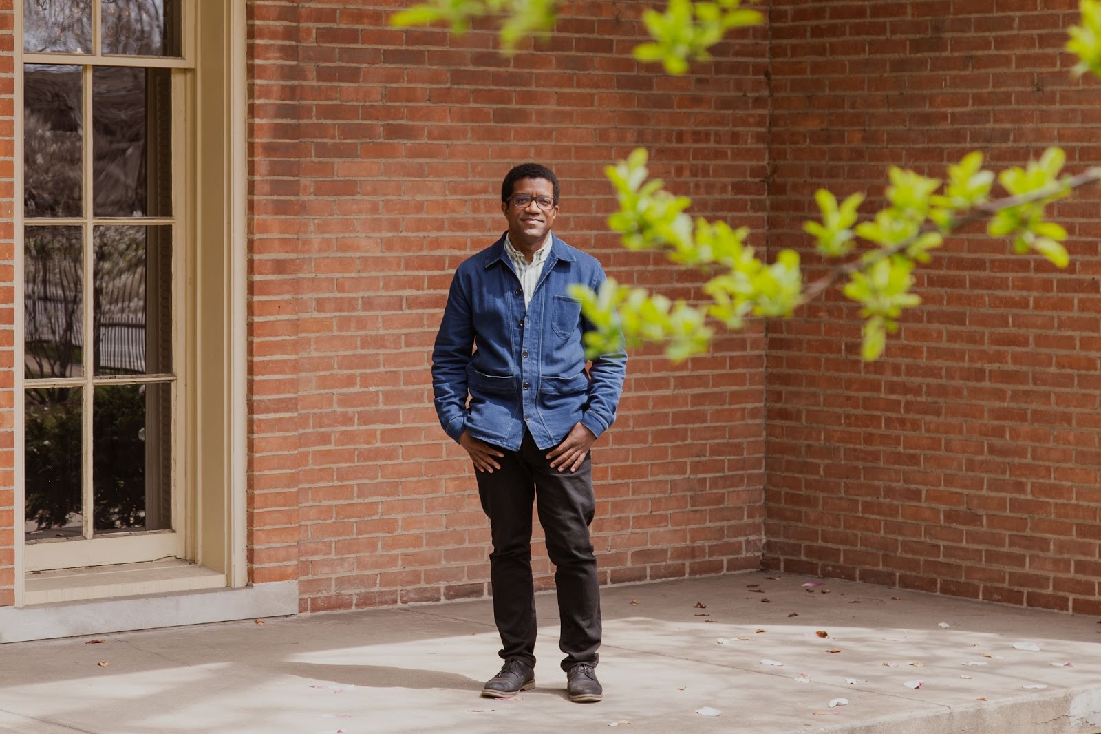 Image: Ross Stanton Jordan stands outside of the Jane Addams Hull House Museum, surrounded by the brick of the building's exterior. There is a tall door to his right and closer to the camera and blurred slightly in the foreground, a couple of tree branches are seen in the top right section of the frame. Ross looks directly into the camera, hands at his pockets. He's wearing a denim jacket, dark brown pants. Photo by Joshua Clay Johnson.