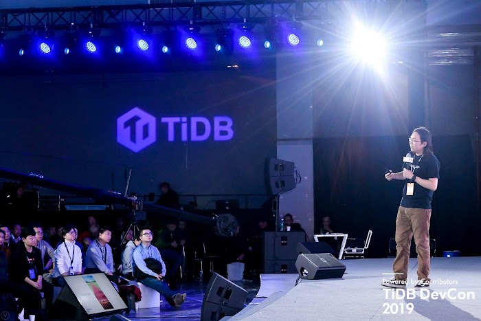 Man presenting on stage in front of audience, with TiBD on wall and 