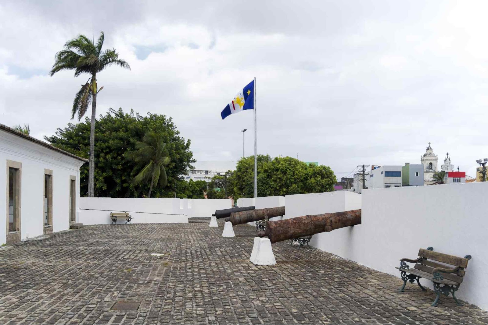 Piso superior do Forte das Cinco Pontas, no Recife Antigo. O pátio com chão de pedra e muros brancos tem três canhões antigos apontados para fora da fortaleza e uma bandeira de Pernambuco.
