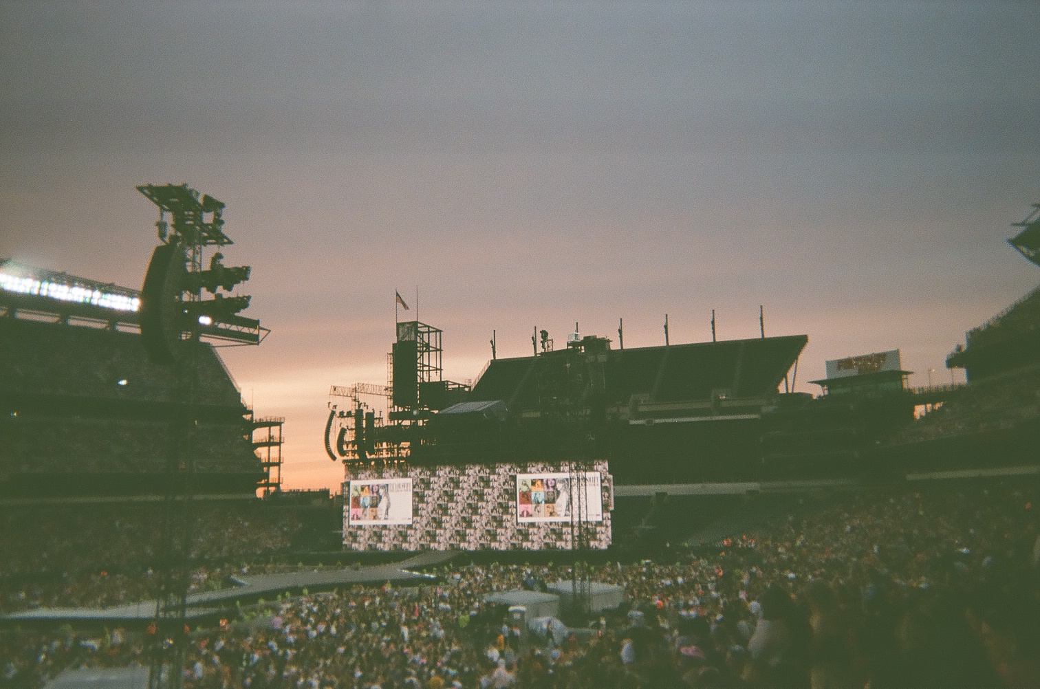 A large stage and a large screen with photos of Taylor Swift. A crowd of people surrounds the stage.