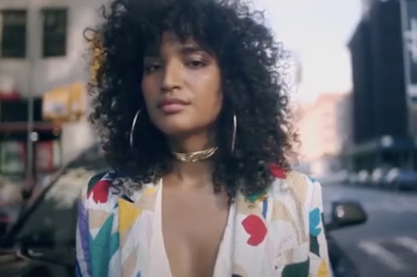 Una mujer trans afrodescendiente con cabello rizado usa joyas de oro y un colorido blazer mientras mira a la cámara