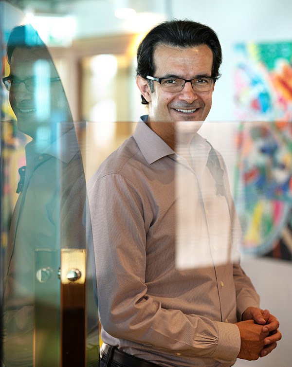 Beshad Behzadi, vice-président du développement de produit de Google Cloud en Suisse, se tient debout en souriant près d'une vitre à travers laquelle le soleil brille et sur laquelle son reflet apparaît. Sur un mur est accroché un grand tableau coloré.