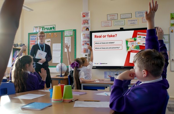 Male teacher stood at the front of a classroom while the students sat down have their hands raised to answer a question.