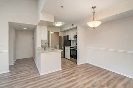 Apartment entry door open to the kitchen, living and dining area with wood plank flooring and neutral walls with white trim throughout 