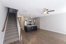 Living room, looking into kitchen, with wood-inspired flooring, a ceiling fan, and carpeted stairs to the second floor