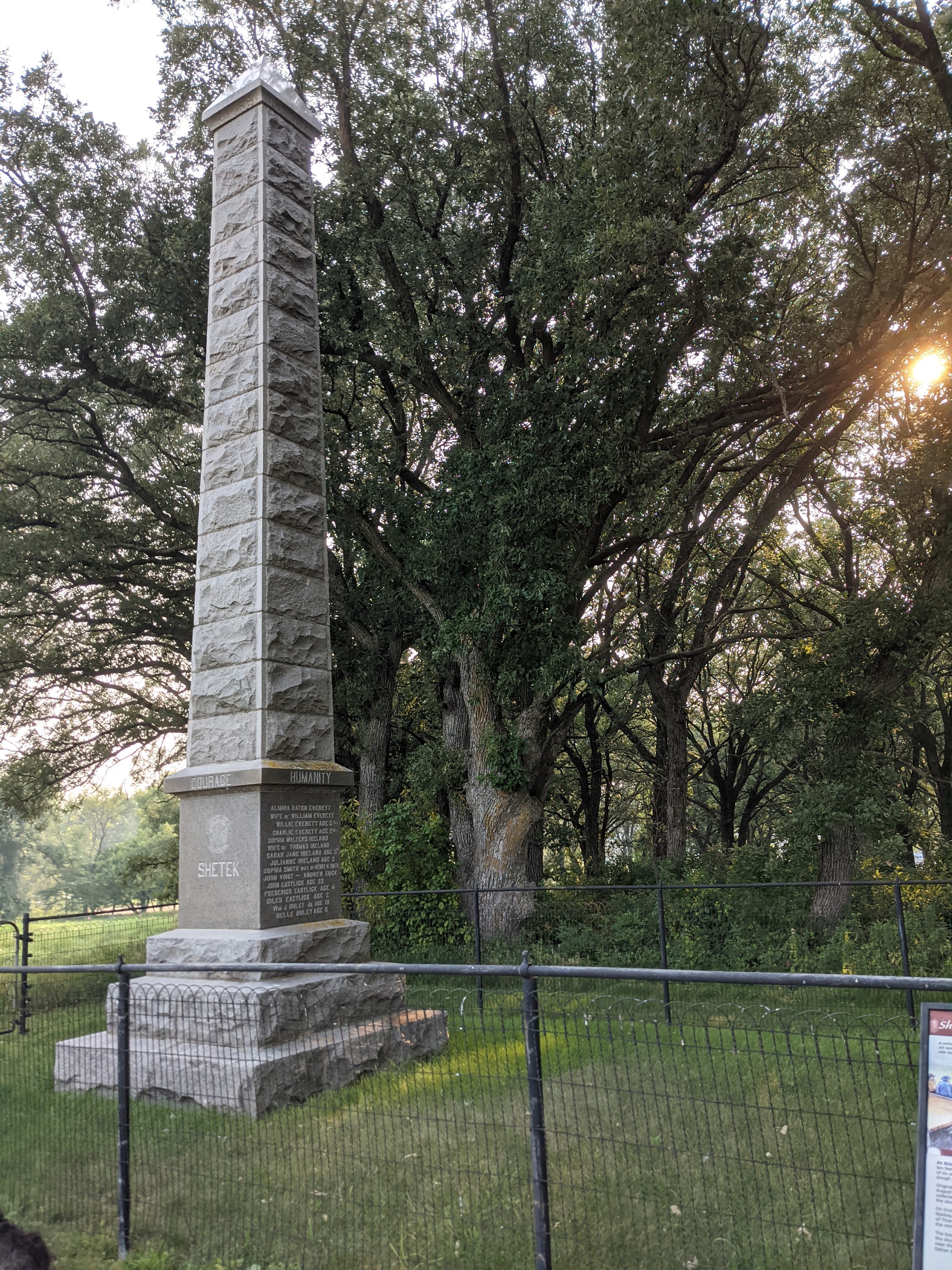 stone oblisk monument surrounded by a small wire fence with trees and the sun in the background