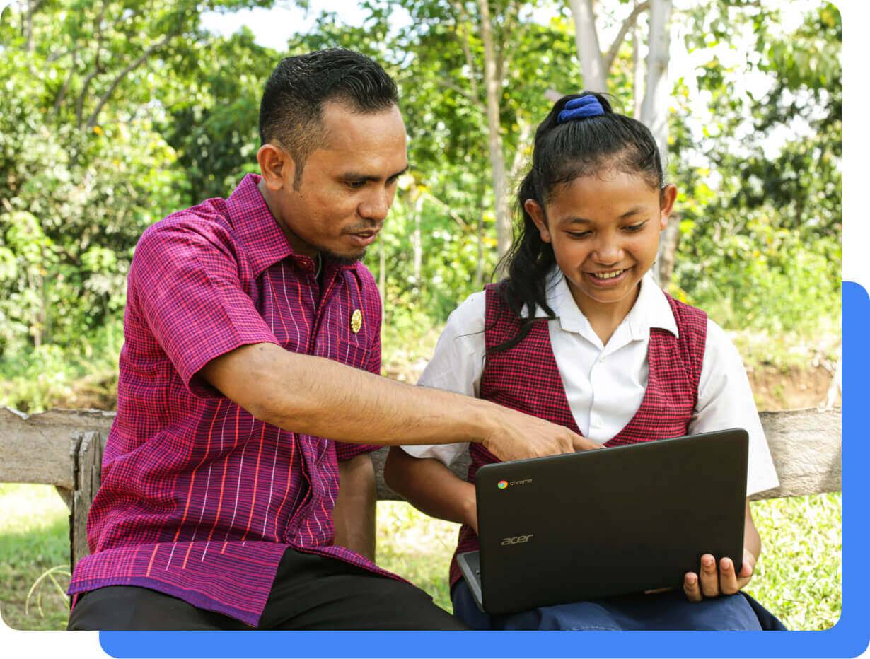 Seorang pengajar laki-laki menunjuk ke arah layar Chromebook seorang siswa perempuan. Mereka berdua sedang duduk bersama di kursi kayu di luar kelas.