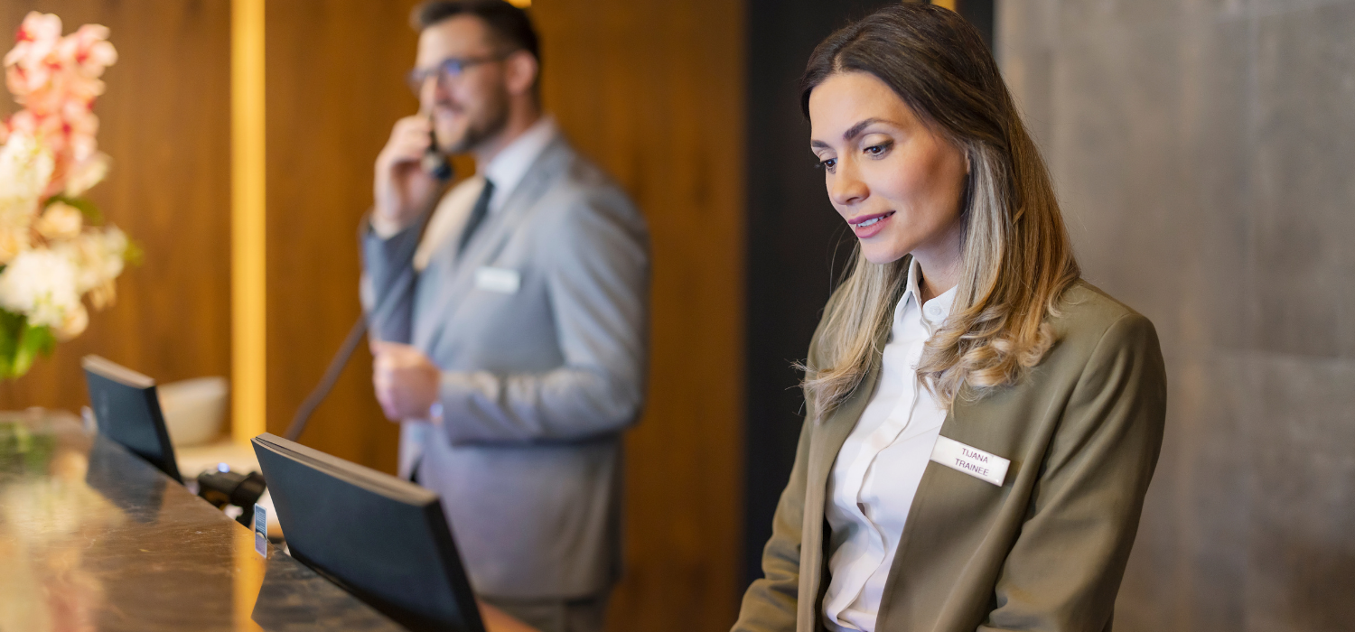 a female receptionist wearing an army green suit with shoulder-length brown hair is managing the hotel guest booking process along with her co-worker who is calling a guest.