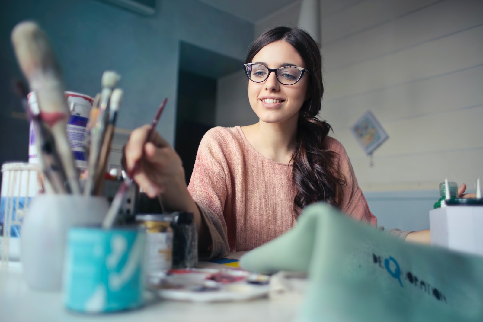 A young girl in glasses is smiling as she dips a paintbrush into a container.  There are paint, brushes, and other supplies on the table.