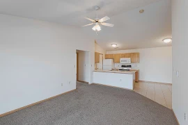 View of carpeted living room, breakfast bar, and tile-floored kitchen, white walls, wood trim, ceiling fan with light